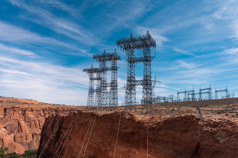 Two tall steel transmission towers on the rim of a red rock canyon, power lines sweeping down across the gorge, with a substation switchyard on the ridge behind them under a blue sky.