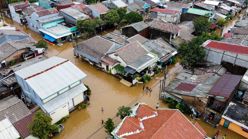 Aerial view of a flooded neighborhood, brown water filling every street between the tiled and metal roofs, with a few people wading along what were the roads.
