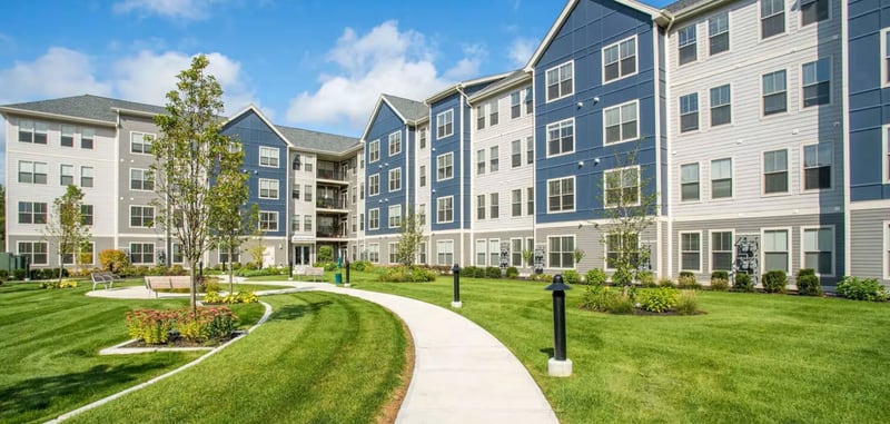 Modern four-story multifamily apartment community with blue and light-gray façades, landscaped courtyard, and pedestrian walkway under a clear sky. The image reflects a stabilizing multifamily market environment, where declining vacancies and renewed rent growth signal early cap rate compression and improving investor confidence, an indicator of shifting dynamics within the broader commercial real estate cycle.