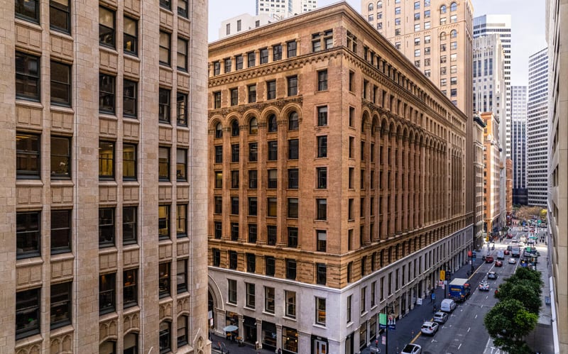 Elevated view of an older brick and terracotta office block with rows of arched windows in downtown San Francisco, hemmed in by glass towers, with traffic on the street below.