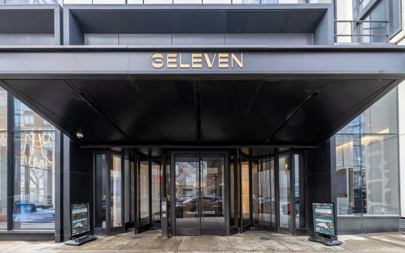 Street-level entrance to 3Eleven, a black canopy carrying the building name in metal letters above revolving glass doors, with a leasing sign board on either side.