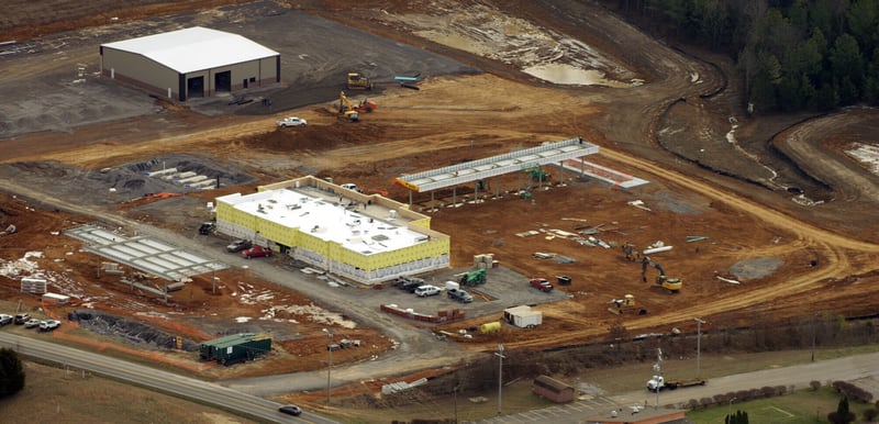 Aerial view of a commercial development under construction, with graded earth, a finished white warehouse, a partly built structure and excavators working across the site.