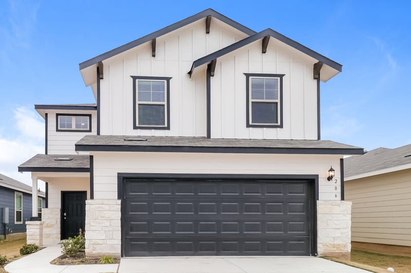 A newly built two story suburban house with white board and batten siding, dark window trim, a wide dark two car garage door and stone accents, under a clear blue sky.