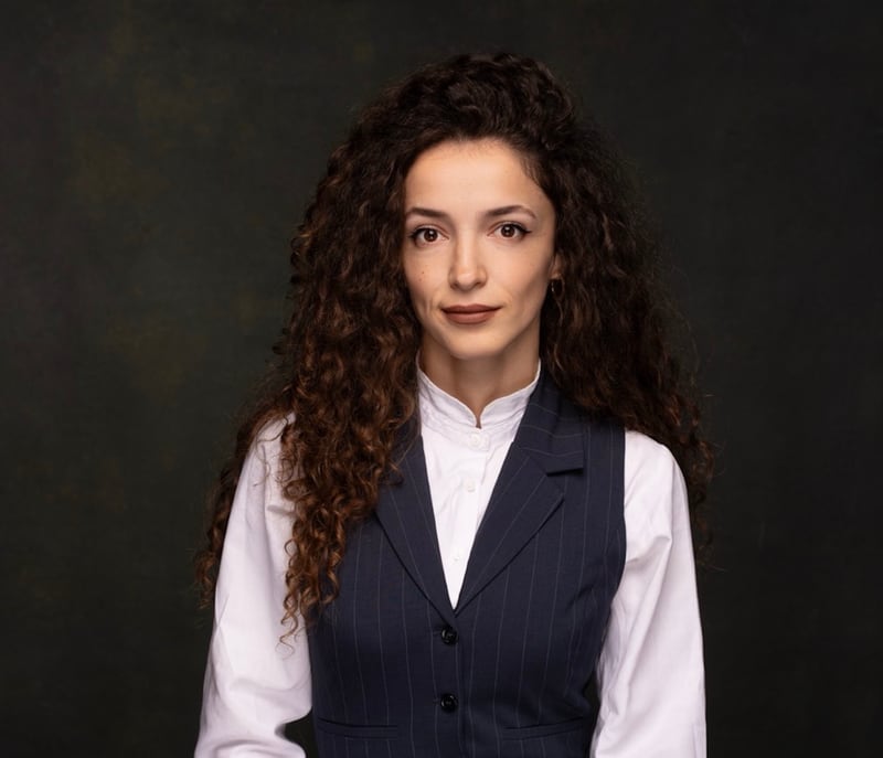 Studio portrait of Manjola Bileri, the analyst credited as the author, wearing a white high-collar blouse under a dark navy pinstripe waistcoat.