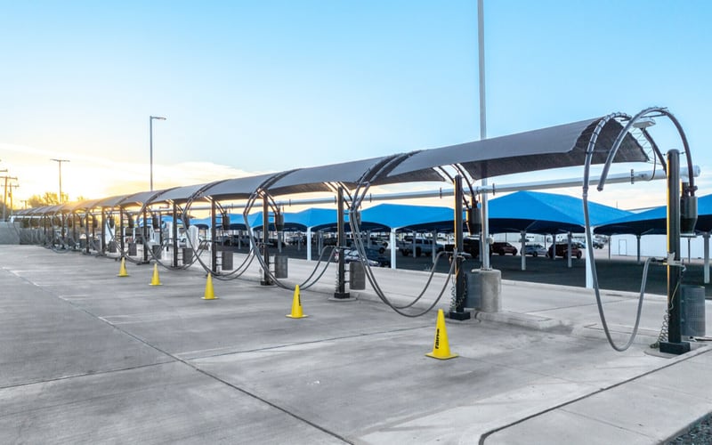 A row of self-serve vacuum stations at a car wash, with looped hose arches under a long canopy, yellow cones on the pavement and blue shade sails over a parking lot behind, the kind of ground-up special-purpose build whose interest reserve the article sizes.