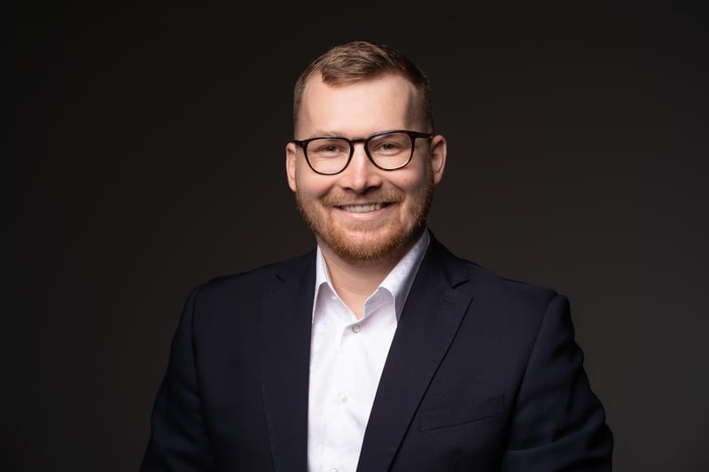 Portrait of Michal Mohelsky, principal of MMCG Invest, a bearded man in dark-rimmed glasses wearing a navy suit and an open-collar white shirt against a dark studio background.