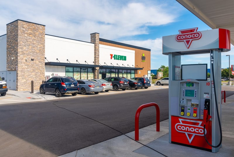 A newly built 7-Eleven convenience store with cars parked along the storefront, viewed from under the fuel canopy beside a Conoco pump island.