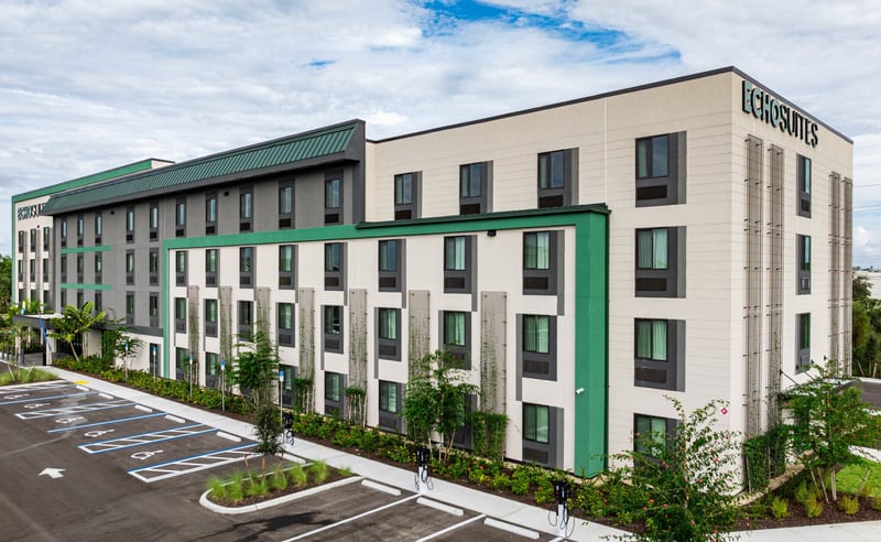 Newly built four-story extended-stay hotel with a green and cream facade and an empty landscaped parking lot in front, photographed under a clear sky.