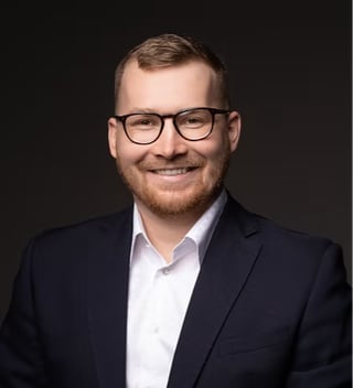 Head and shoulders portrait of Michal Mohelsky, principal of MMCG Invest, in dark-rimmed glasses, a navy suit and an open-collar white shirt against a dark studio background.
