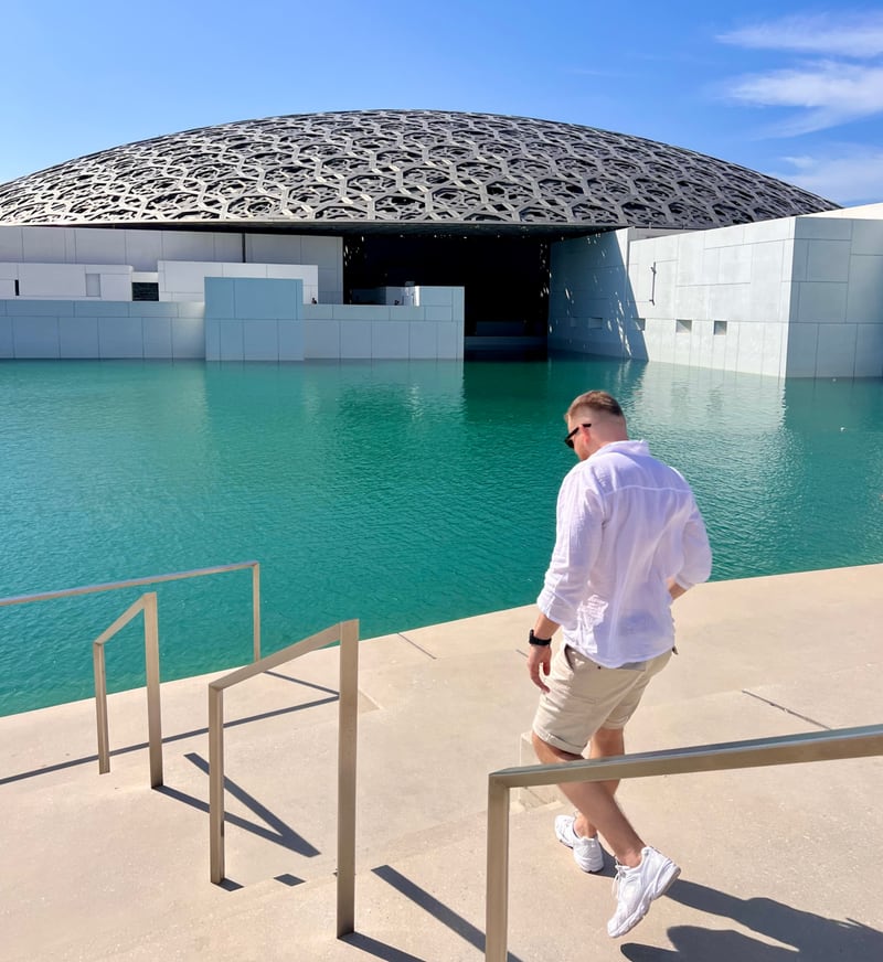 The latticed dome of the Louvre Abu Dhabi on Saadiyat Island rises over turquoise water while a visitor in a white shirt walks along the waterfront railing.