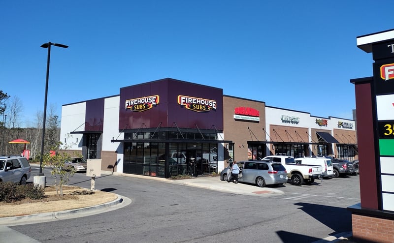 A Firehouse Subs restaurant on the corner of a suburban strip center, with two lit signs on a dark facade, neighboring storefronts and cars parked out front.