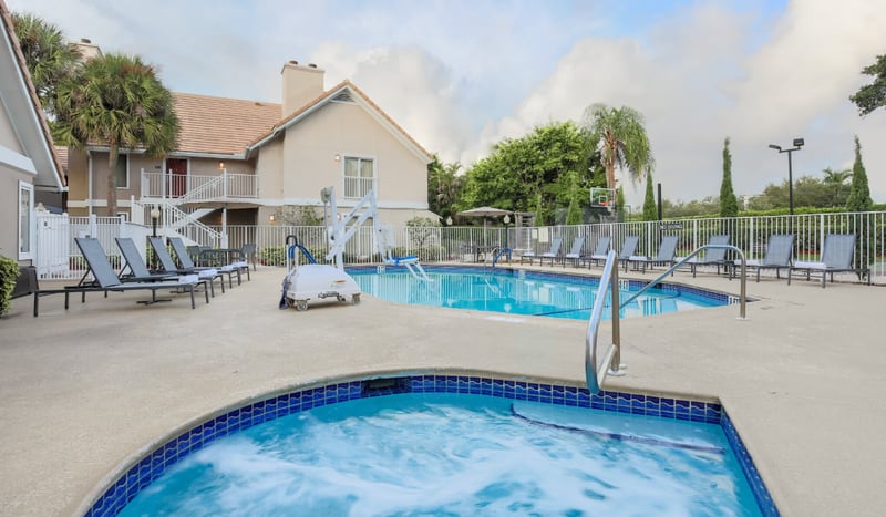 Outdoor pool area at the Residence Inn Boca Raton, with a bubbling whirlpool in the foreground, lounge chairs and a pool lift beside the main pool, and the two-story residential-style buildings behind.