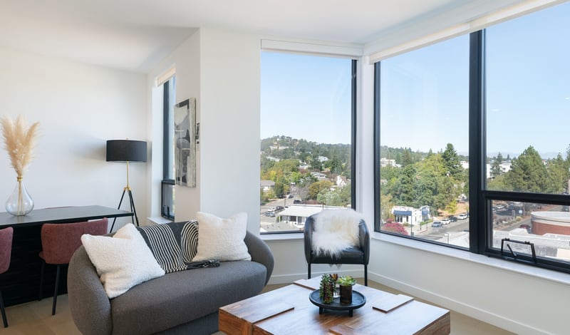 A furnished apartment living room with a grey sofa, an armchair and a wood coffee table beside floor-to-ceiling corner windows that look out over trees, houses and wooded hills.