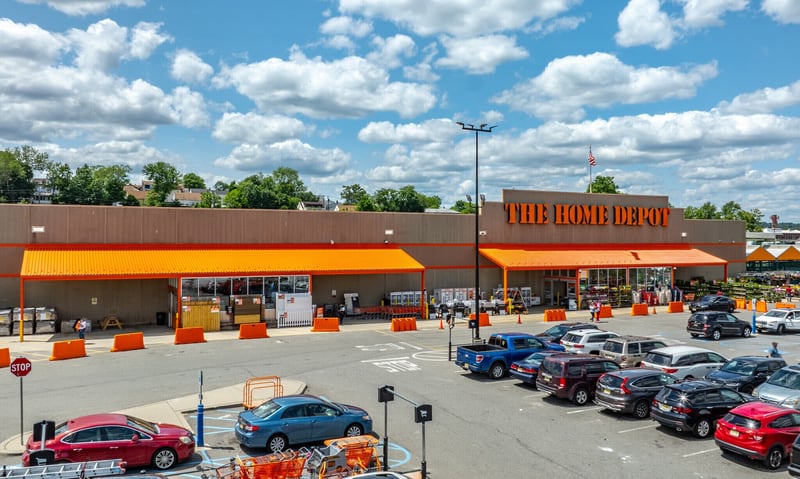 Exterior of a Home Depot big-box store on a sunny day, its orange entrance canopies and store sign facing a parking lot full of cars, with the garden center at the far end.