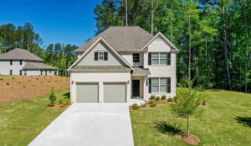 A new two-story suburban single-family house with light brick and siding, a double garage and a concrete driveway on a fresh lawn, with pine woods behind it.