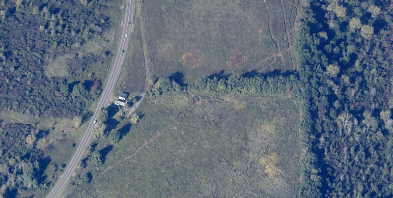 Aerial view of the proposed site at 5171 State Route 31 in the Town of Clay: open, formerly agricultural fields with scattered brush, a single small structure at the road frontage, and mature woodland along the northern and eastern edges.