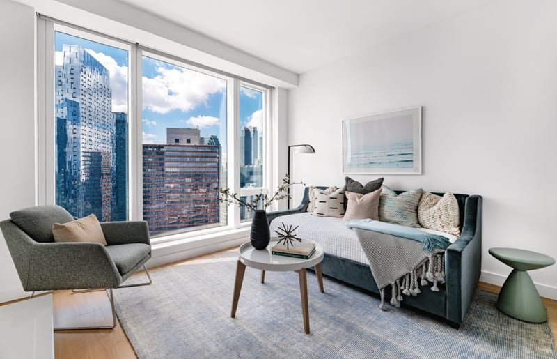 A furnished living room in one of Sven's premium units, with floor-to-ceiling windows facing neighbouring high-rises, a grey sofa, an armchair and a round coffee table on a blue rug.