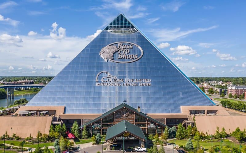 The Bass Pro Shops Pyramid in Memphis, a glass-clad pyramid carrying the Bass Pro Shops logo and Ducks Unlimited Waterfowling Heritage Center lettering above a timber lodge-style entrance, with the river and a bridge behind it.