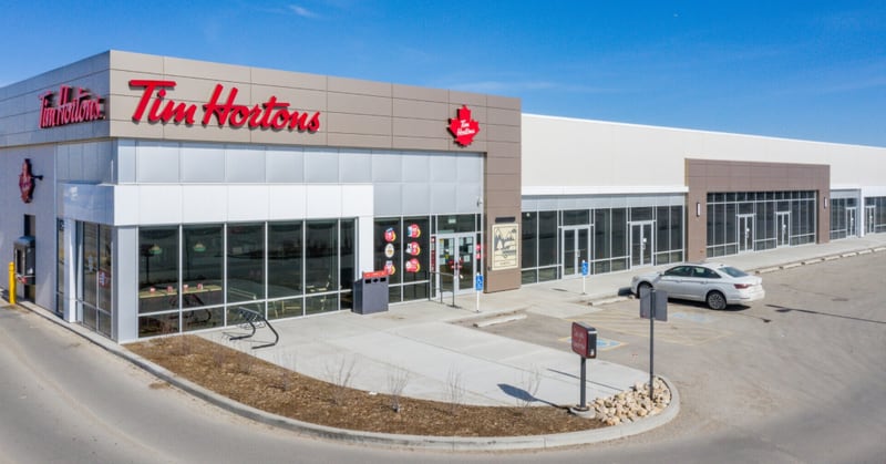 A Tim Hortons cafe at the end of a new single-storey retail strip, its red script sign and maple-leaf logo on a grey and white facade with one car parked outside.