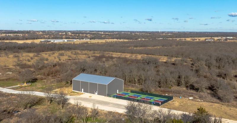 Aerial view of a grey steel building beside a bank of outdoor pickleball courts in blue, green and red, set in bare winter woodland with open land beyond.