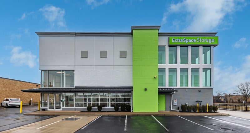 A two-story Extra Space Storage facility with a white facade, a bright green vertical accent and green sign, a glass-walled office at ground level and an empty parking lot in front under an overcast sky.
