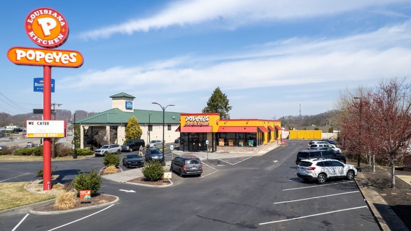 A Popeyes Louisiana Kitchen restaurant in red and yellow with its tall orange roadside sign in the foreground, cars parked out front and a hotel building behind it.