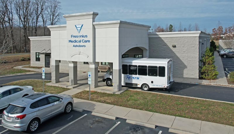 Fresenius Medical Care dialysis clinic in Asheboro: a single-storey tan building with the company's sign on a covered entrance portico, a white patient transport shuttle bus parked under the canopy and cars in the lot in front.