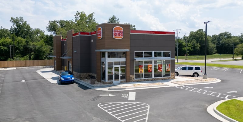 A newly built Burger King restaurant with a dark brown and grey facade, red logo signs and a drive-thru lane, seen across an empty parking lot with a single car at the window.
