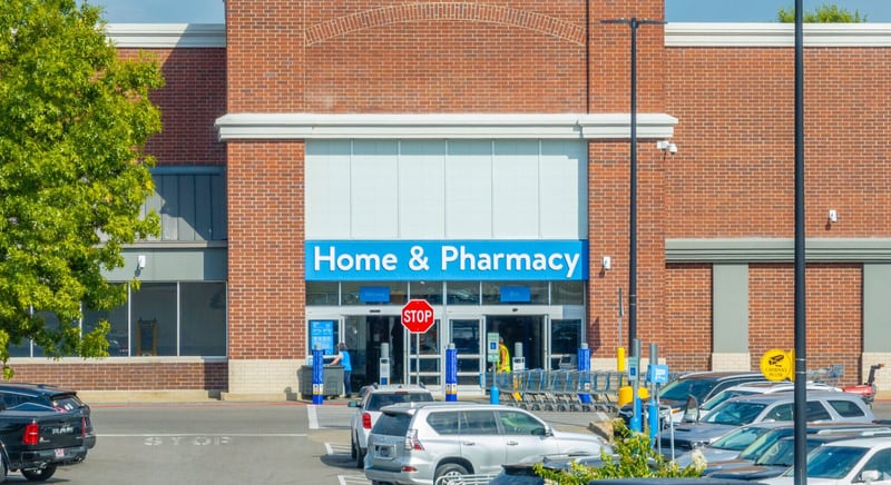 A Walmart store's brick entrance under a blue Home & Pharmacy sign, with shopping carts, a stop sign and a parking lot of cars in front.