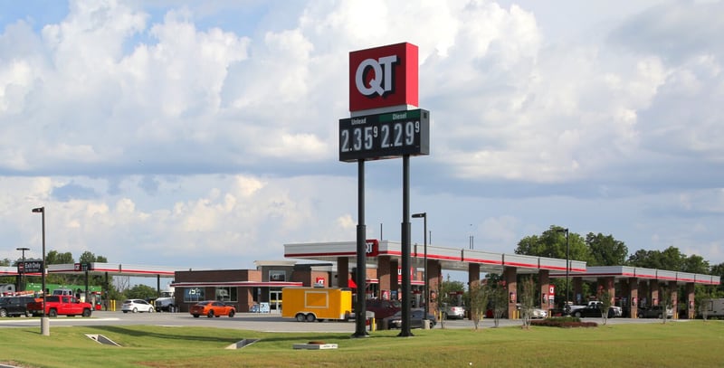 QuikTrip gas station exterior with a tall red QT price sign reading $2.35 and $2.29 above a wide fuel canopy and convenience store, with cars and a yellow truck in the lot under a cloudy sky.