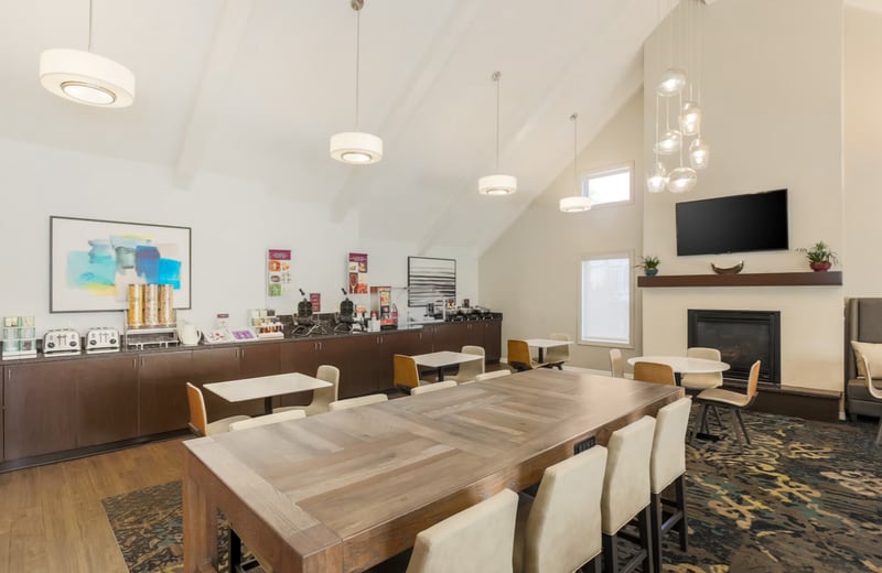 Breakfast and lounge room at the Residence Inn Boca Raton with a long communal wood table, a buffet counter with coffee stations, and a fireplace under a wall-mounted television.