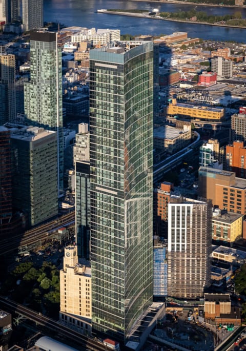Sven, the 70-story glass apartment tower at 2959 Northern Blvd, seen from the air rising above lower Long Island City buildings, with the East River behind it and an older clock-tower building at its base.