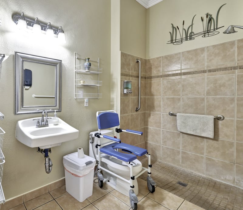 An accessible care facility bathroom with a roll-in tiled shower, wall grab bars, a wheeled blue shower and commode chair, and a wall-mounted sink.