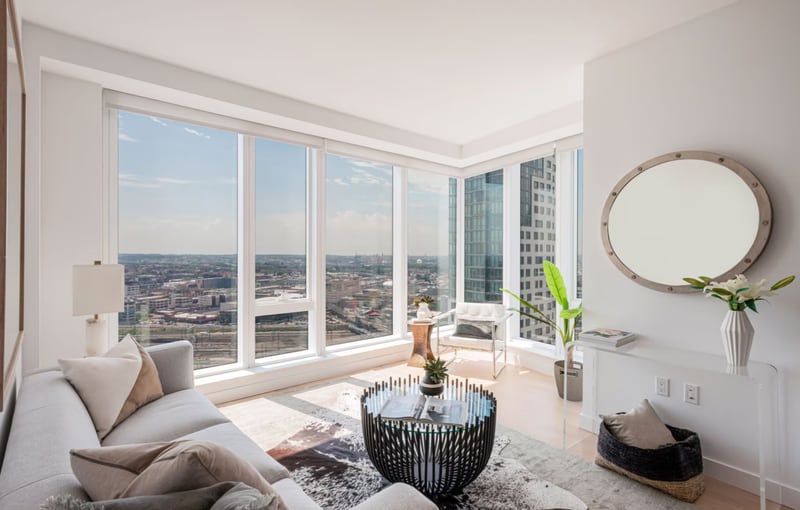 A corner living room in a high rise apartment, pale sectional sofa and round mirror, floor to ceiling windows on two walls looking out over Long Island City rooftops and towers.