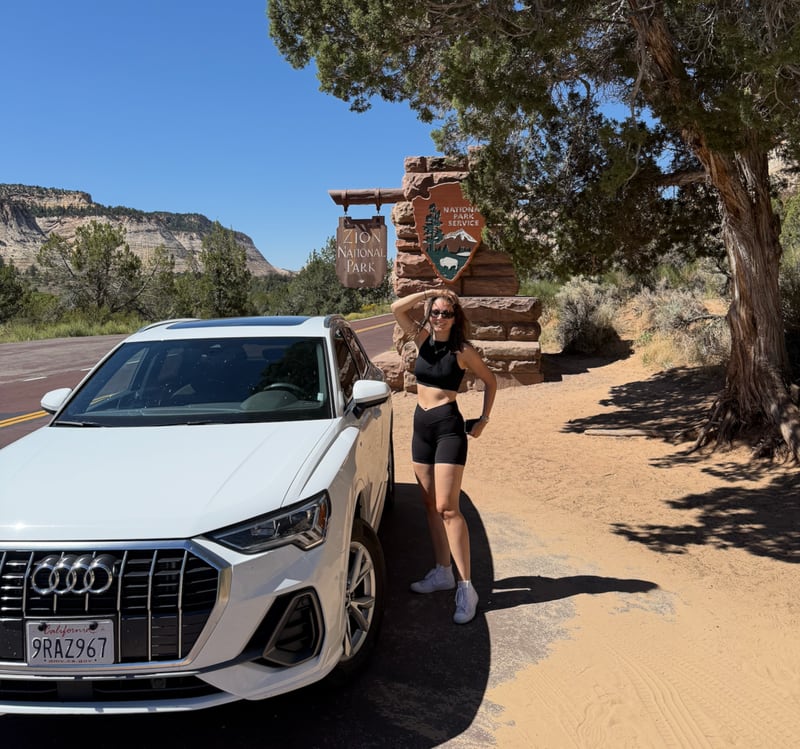 A woman stands beside a white SUV at the stone and timber entrance sign for Zion National Park, with red rock cliffs and desert scrub behind her.