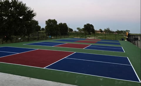 Newly surfaced outdoor pickleball courts with blue and red playing surfaces and white lines on a green surround, fenced and set beside open ground at dusk.