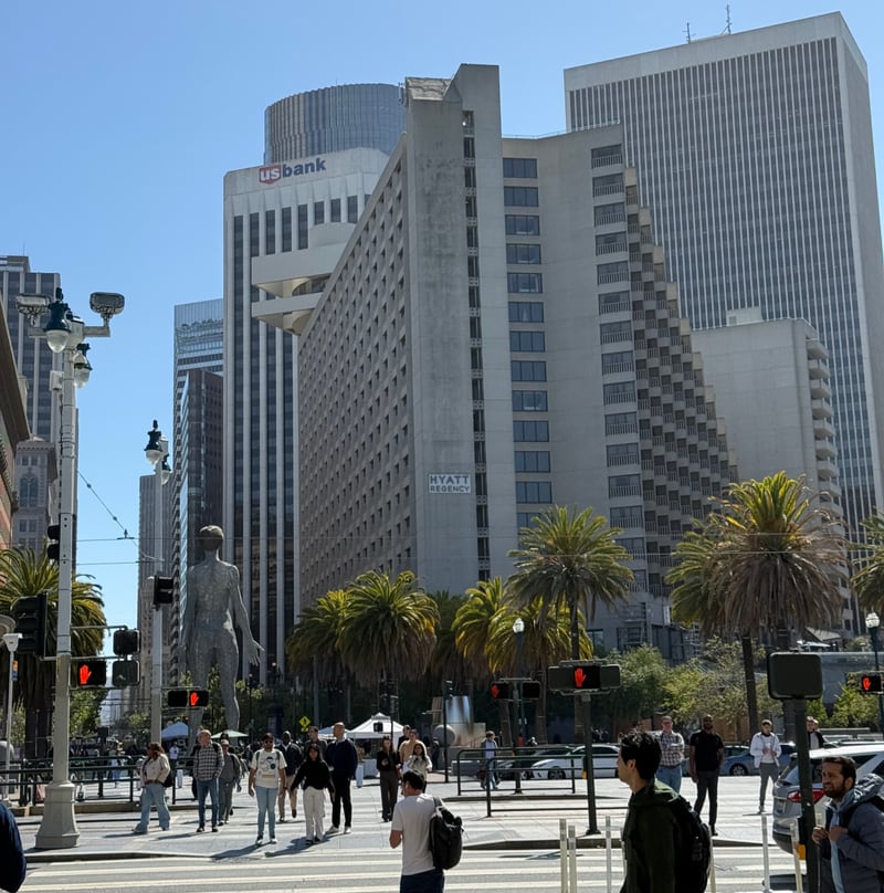 Pedestrians crossing a downtown San Francisco intersection at the foot of Market Street on a clear day, with the stepped concrete Hyatt Regency, the US Bank tower, palm trees and a tall steel figure sculpture in the plaza behind them.