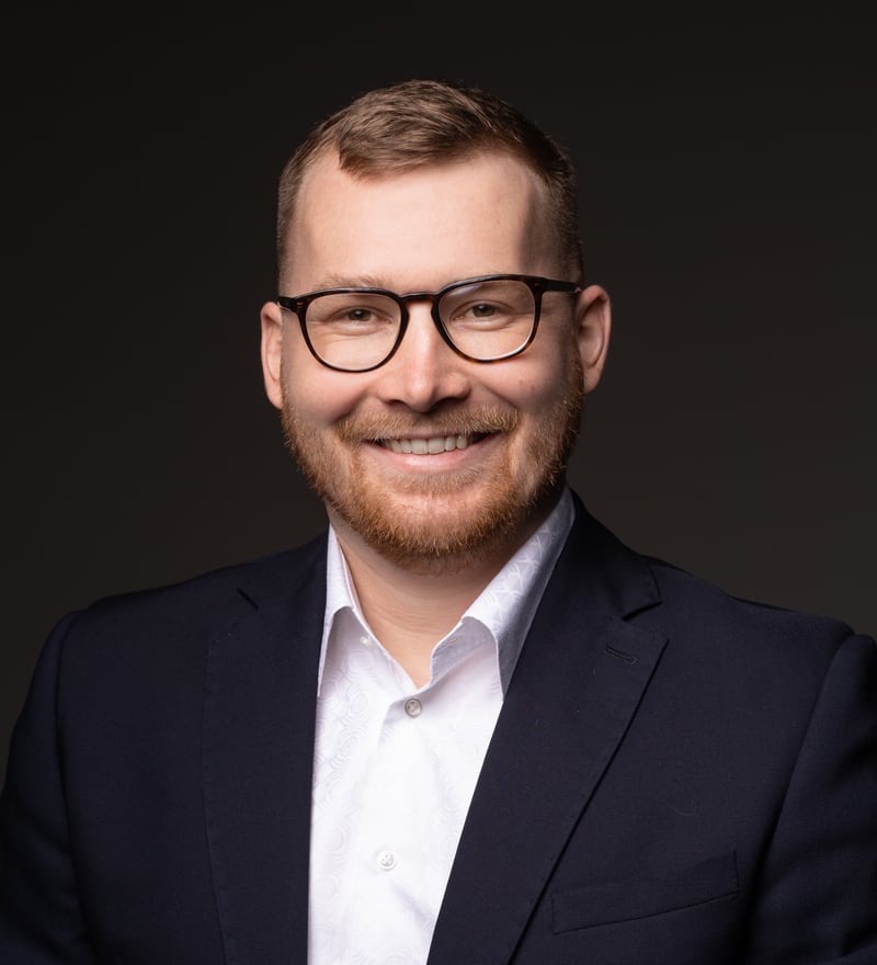 Michal Mohelsky, J.D., Principal of MMCG Invest, photographed from the chest up in a dark navy suit and open-collar white shirt against a near black background.